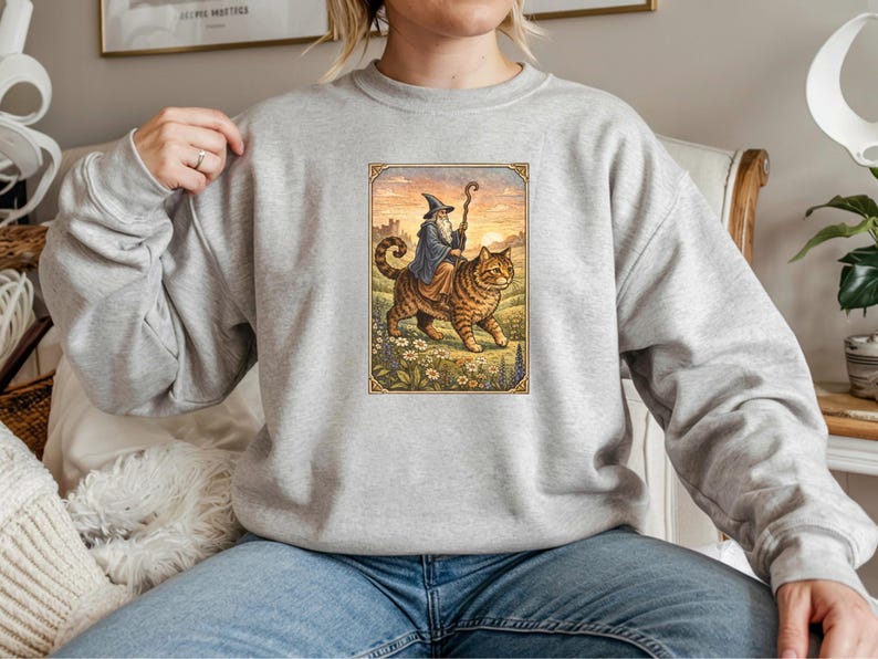 May include: A light gray sweatshirt featuring a whimsical illustration of a wizard riding a large tabby cat. The wizard wears a blue robe and hat, holding a staff. The cat walks through a field of flowers.