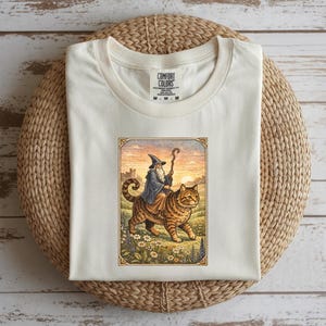 May include: Cream-colored t-shirt featuring a whimsical illustration of a wizard riding a large tabby cat. The wizard wears a blue robe and a pointed hat, holding a staff. The cat walks through a field with flowers.