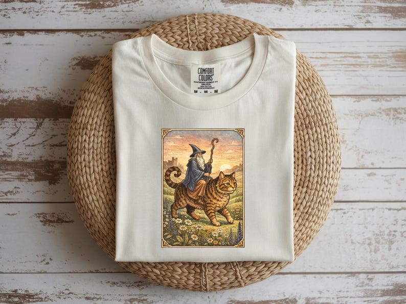May include: Cream-colored t-shirt featuring a whimsical illustration of a wizard riding a large tabby cat. The wizard wears a blue robe and a pointed hat, holding a staff. The cat walks through a field with flowers.