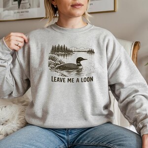 May include: A light gray sweatshirt featuring a black and white illustration of a loon on a lake with trees. The text "LEAVE ME A LOON" is printed below the image. The sweatshirt is a casual style.