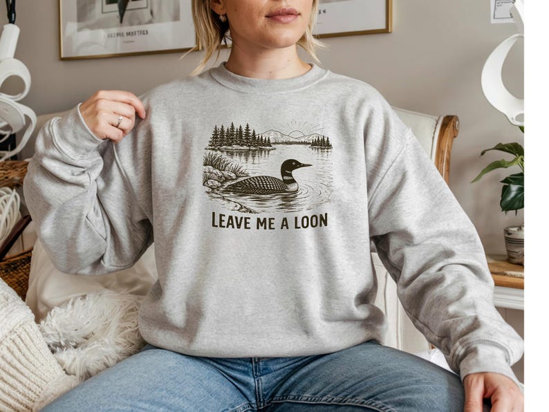 May include: A light gray sweatshirt featuring a black and white illustration of a loon on a lake with trees. The text "LEAVE ME A LOON" is printed below the image. The sweatshirt is a casual style.