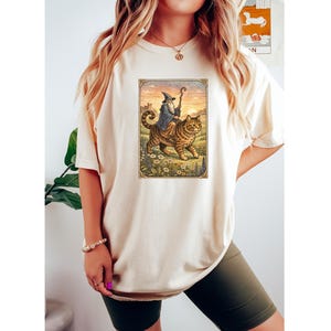 May include: Cream-colored t-shirt featuring a framed illustration of a wizard riding a large tabby cat through a field of flowers. The wizard wears a blue robe and holds a staff. The background shows a castle and a sunset.