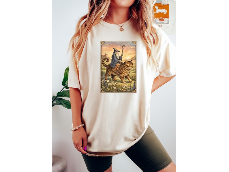 May include: Cream-colored t-shirt featuring a framed illustration of a wizard riding a large tabby cat through a field of flowers. The wizard wears a blue robe and holds a staff. The background shows a castle and a sunset.