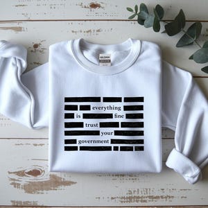 May include: White crewneck sweatshirt with the text "everything is fine trust your government" in a black, blocky font. The sweatshirt is made of a soft material and has long sleeves.