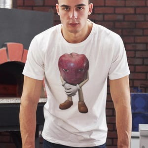May include: A man wearing a white t-shirt with a red apple graphic. The apple has a face and is wearing shoes. He is holding a pizza with pepperoni on a wooden board.