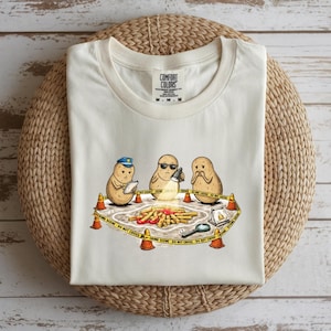 May include: Off-white t-shirt with a comical design of three potato characters at a crime scene. The scene includes crime scene tape, cones, and a magnifying glass. The shirt has a crew neck and the brand name "Comfort Colors".