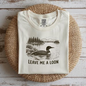 May include: Cream-colored t-shirt featuring a black and white illustration of a loon swimming in a lake, with trees and mountains in the background. The text "LEAVE ME A LOON" is printed below the image. The shirt is folded on a woven mat.