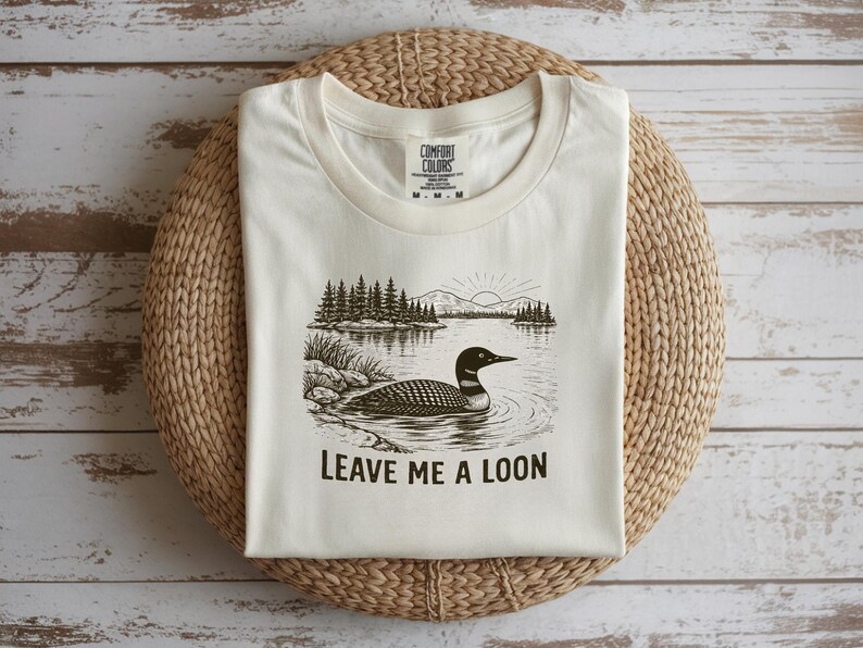 May include: Cream-colored t-shirt featuring a black and white illustration of a loon swimming in a lake, with trees and mountains in the background. The text "LEAVE ME A LOON" is printed below the image. The shirt is folded on a woven mat.