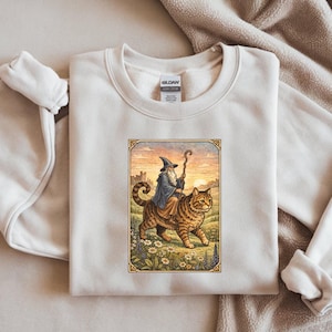 May include: Cream-colored sweatshirt featuring a framed illustration of a wizard riding a large tabby cat. The wizard wears a blue robe and hat, holding a staff. The cat walks through a field of flowers, with a castle in the background.