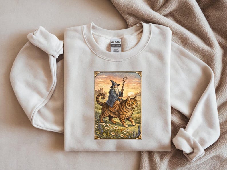 May include: Cream-colored sweatshirt featuring a framed illustration of a wizard riding a large tabby cat. The wizard wears a blue robe and hat, holding a staff. The cat walks through a field of flowers, with a castle in the background.