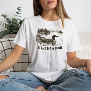 May include: White t-shirt featuring a black and white graphic of a loon swimming in a lake with trees and mountains. The text "LEAVE ME A LOON" is printed below the graphic. The shirt is worn by a person.