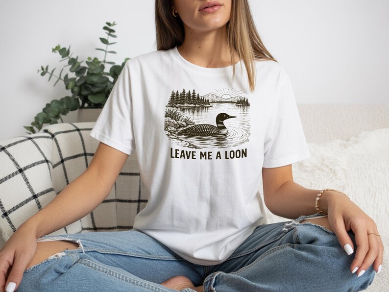May include: White t-shirt featuring a black and white graphic of a loon swimming in a lake with trees and mountains. The text "LEAVE ME A LOON" is printed below the graphic. The shirt is worn by a person.