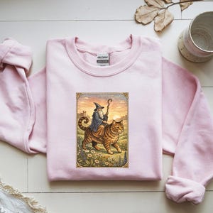 May include: A light pink Gildan sweatshirt featuring a framed illustration of a wizard riding a large tabby cat. The wizard wears a blue robe and hat, holding a staff. The cat walks through a field of flowers.