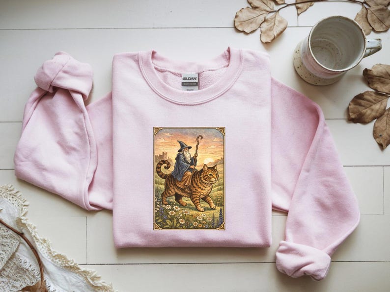 May include: A light pink Gildan sweatshirt featuring a framed illustration of a wizard riding a large tabby cat. The wizard wears a blue robe and hat, holding a staff. The cat walks through a field of flowers.