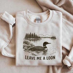 May include: Cream-colored sweatshirt with a graphic of a loon swimming in a lake, with trees and mountains in the background. The text "LEAVE ME A LOON" is printed below the graphic. The sweatshirt is folded.
