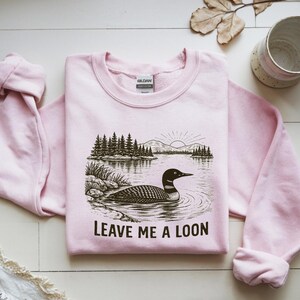May include: A light pink sweatshirt with a graphic of a loon swimming in a lake, with trees and a rising sun in the background. The text "LEAVE ME A LOON" is below the image. The sweatshirt is made of a soft material.