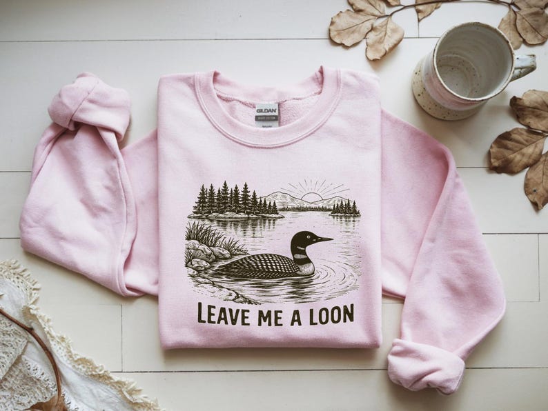 May include: A light pink sweatshirt with a graphic of a loon swimming in a lake, with trees and a rising sun in the background. The text "LEAVE ME A LOON" is below the image. The sweatshirt is made of a soft material.