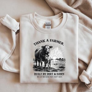 May include: Cream-colored sweatshirt with a black and white graphic of a cow and calf, with the text "THANK A FARMER." Below the image, the text reads "BUILT BY DIRT & DAWN" and "WE EAT BECAUSE THEY DON'T QUIT."
