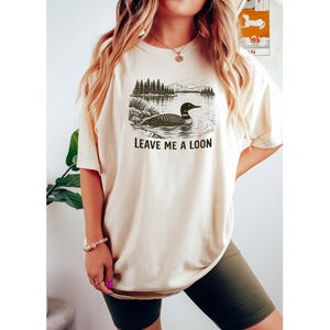 May include: Cream-colored t-shirt featuring a black and white illustration of a loon on a lake with trees and mountains. The text "LEAVE ME A LOON" is printed below the image. The shirt is a casual, unisex style.