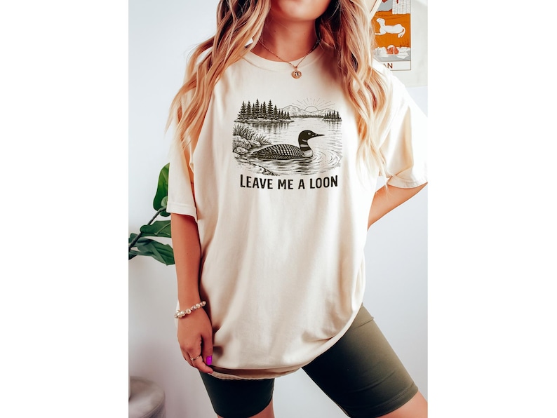 May include: Cream-colored t-shirt featuring a black and white illustration of a loon on a lake with trees and mountains. The text "LEAVE ME A LOON" is printed below the image. The shirt is a casual, unisex style.