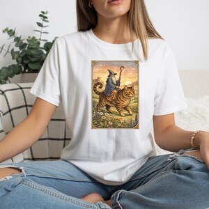 May include: White t-shirt featuring a colorful illustration of a wizard riding a large tabby cat. The wizard wears a blue robe and pointed hat, holding a staff. The cat walks through a field of flowers.
