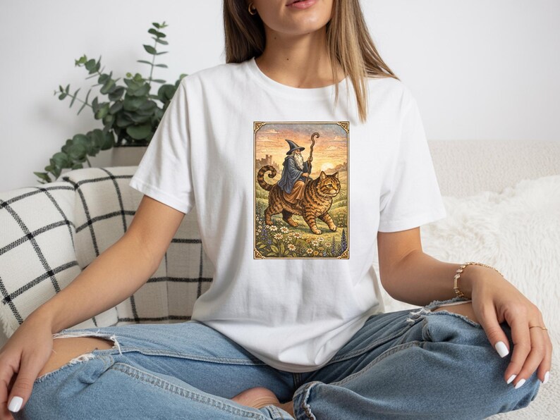 May include: White t-shirt featuring a colorful illustration of a wizard riding a large tabby cat. The wizard wears a blue robe and pointed hat, holding a staff. The cat walks through a field of flowers.