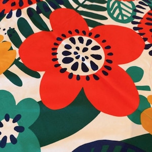 Op de afbeelding: Een textielstof met een opvallend bloemenpatroon van grote rode, blauwgroene en gele bloemen op een witte achtergrond. Het ontwerp bevat donkerblauwe accenten in de bloemharten en donkergroene bladeren die over de hele compositie zijn verspreid.