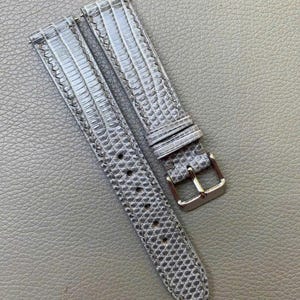 May include: A two-piece gray watch strap featuring a textured, reptilian-inspired scale pattern. The strap includes a polished silver-tone metal buckle, a matching sliding keeper loop, and a series of punched holes for adjustable sizing.