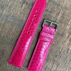 Può includere: Un cinturino per orologio rosa acceso caratterizzato da una trama testurizzata simile a scaglie su tutta la superficie. Il cinturino è composto da due parti, una con una serie di fori e l'altra con una fibbia in metallo argentato e due passanti.