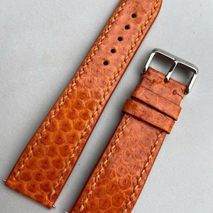 May include: A two-piece watch strap in a vibrant orange hue with a distinct hexagonal scale texture. The strap features light-colored stitching along the edges, a silver-toned metal buckle, and two matching leather keepers for secure fastening.