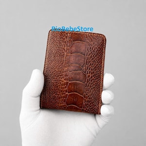 Puede incluir: Una cartera plegable marrón con un patrón de cuero de pata de avestruz texturizado y un diseño central distintivo. El texto RioBebeStore aparece en azul en la parte superior. La superficie tiene un acabado brillante con detalles de escamas.