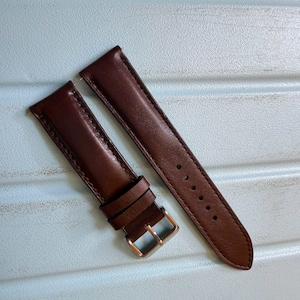 Puede incluir: Una correa de reloj de cuero marrón oscuro de dos piezas con costuras lisas en los bordes, dos trabillas de cuero y una hebilla de metal plateado pulido. La pieza más larga incluye una serie de seis orificios circulares de ajuste.