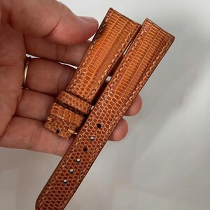 Può includere: Un cinturino per orologio in due pezzi di colore marrone medio con un motivo testurizzato ispirato alle squame di rettile. Il cinturino presenta cuciture visibili lungo i bordi, un passante fisso e una serie di fori per la chiusura.