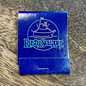 May include: A blue matchbook with a white outline of a boat and the text "Rusty Scupper" in white with a blue outline. The matchbook has a blue background with a white stripe.