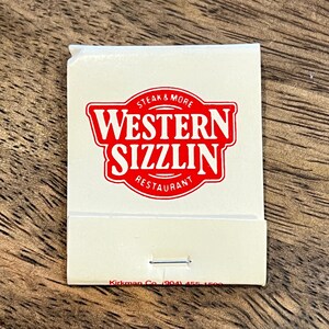 May include: A white matchbook with a red logo that says "Steak & More Western Sizzlin Restaurant" on the front. The matchbook has a silver metal strip on the back.
