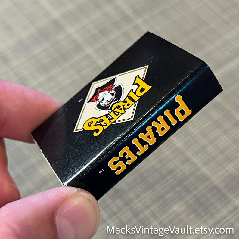 May include: A black box featuring the Pittsburgh Pirates logo in yellow and white. The word "PIRATES" is printed in yellow on the side of the box. The logo depicts a pirate wearing a red hat.