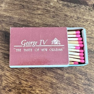 May include: A red matchbox with the text "George IV" and "The Taste of New Orleans" printed on the front. The matchbox is open and shows a stack of wooden matches with pink tips.