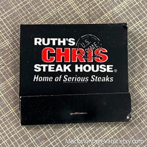 May include: A black matchbook from Ruth's Chris Steak House. The matchbook features the restaurant's name in white and red lettering, with the words "Home of Serious Steaks" below. A U.S. Prime stamp is also visible.