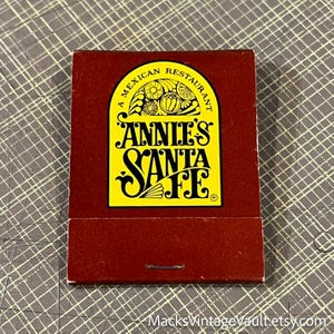 May include: A vintage matchbook from "Annie's Santa Fe," a Mexican restaurant. The matchbook has a dark red cover with a yellow and black logo. The logo features the text "Annie's Santa Fe" and "A Mexican Restaurant."