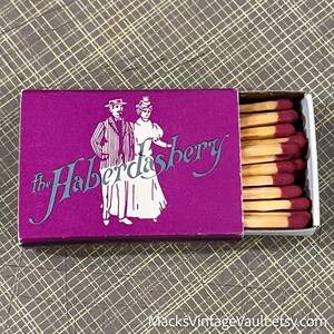 May include: A vintage matchbox with a purple cover featuring the text "the Haberdashery" and illustrations of a man and woman in period clothing. The matchbox is open, revealing several matches with red tips.