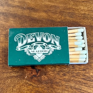May include: A green matchbook with the text "Devon Seafood Grill" in white lettering. The matchbook is open and shows a stack of matches with white tips.