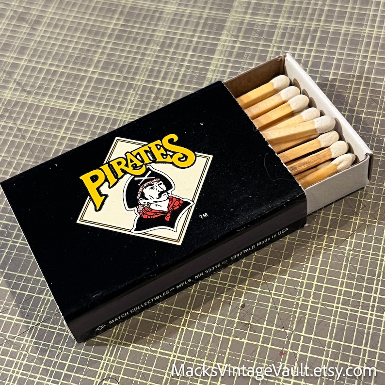 May include: Black matchbox with the word "Pirates" in yellow and a pirate logo. The matchbox is open, revealing wooden matches with white tips. The text "MATCH COLLECTIBLES MPLS, MN 55416 1992 MLB Made in USA" is printed on the box.