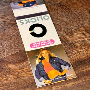 May include: A matchbook cover with a black and blue circle logo and the text "OLICKS" in white. The text "WE'RE SERIOUS ABOUT THE GAME" is in pink. A woman in a denim jacket and pink tank top is pictured on the back of the matchbook.