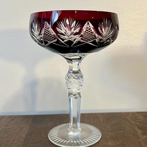 May include: A clear crystal wine glass with a ruby red bowl. The bowl features a cut design of geometric shapes and leaf-like patterns. The stem has a decorative ball and a clear, round base.