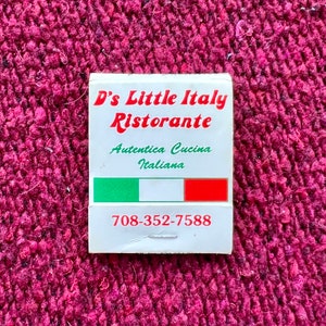 May include: A white matchbook with red and green accents. The text on the matchbook reads "D's Little Italy Ristorante" and "Autentica Cucina Italiana". The phone number 708-352-7588 is printed on the matchbook.