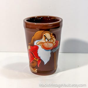 May include: A brown ceramic tumbler featuring a colorful graphic of the character Grumpy from Snow White and the Seven Dwarfs. The character has a white beard, red clothing, and a brown hat, leaning on a pickaxe. The text MacksVintageVault.etsy.com is visible.