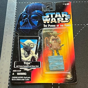 May include: A Star Wars "The Power of the Force" action figure of Yoda in a plastic package. The package features Darth Vader's helmet and the text "Yoda with Jedi Trainer Backpack and Gimer Stick!" Ages 4 & Up. Warning: Choking Hazard.