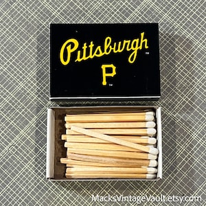 May include: A box of matches with the word "Pittsburgh" in yellow script and a yellow "P" on a black background. The matchbox is open, revealing a collection of wooden matches with white tips.