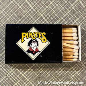 May include: A black matchbox with the word "PIRATES" in yellow lettering and a pirate illustration. The matchbox is open, revealing a row of wooden matches with white tips.