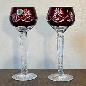 May include: Two clear glass wine glasses with a deep red, cut-glass bowl. The glasses have a long, clear, faceted stem and a clear, faceted base. The bowl of one glass has a sticker that reads "Made in Poland".